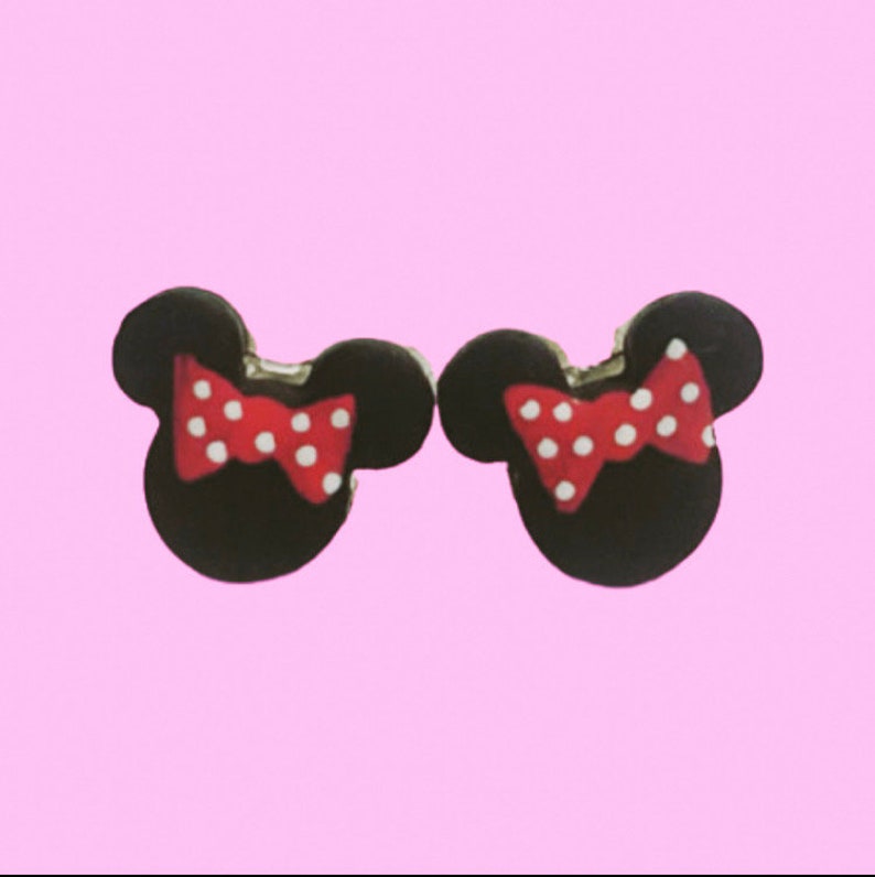May include: A pair of black mouse-shaped earrings with red bows and white polka dots. The earrings are set against a light pink background.