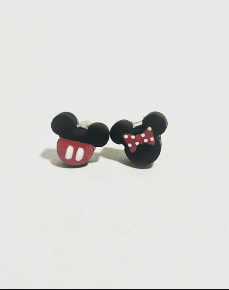 May include: A pair of black and red Mickey Mouse and Minnie Mouse stud earrings. The Mickey Mouse earring has a red base with white oval details. The Minnie Mouse earring has a red and white polka dot bow.