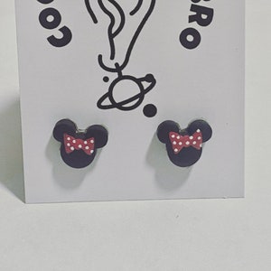 May include: A pair of black stud earrings in the shape of a mouse head, each with a red bow adorned with white polka dots. The earrings are presented on a white card with a black illustration and text.
