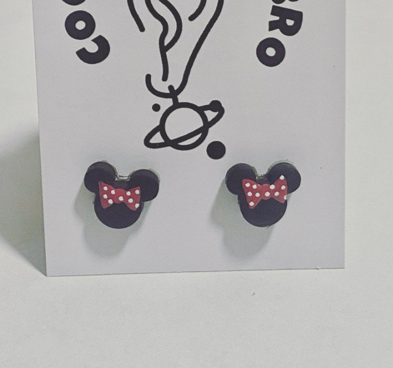 May include: A pair of black stud earrings in the shape of a mouse head, each with a red bow adorned with white polka dots. The earrings are presented on a white card with a black illustration and text.