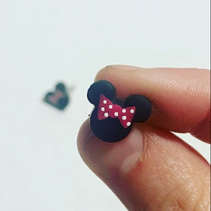 May include: A black mouse head stud earring with a red bow and white polka dots. The earring is held between fingers against a white backdrop. Another earring is slightly out of focus in the background.