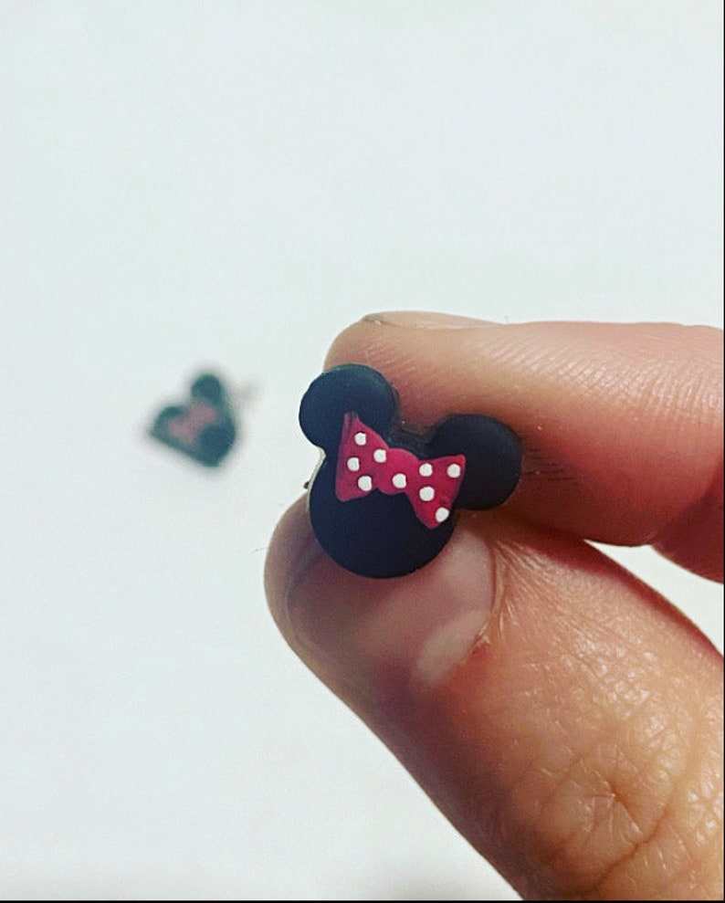 May include: A black mouse head stud earring with a red bow and white polka dots. The earring is held between fingers against a white backdrop. Another earring is slightly out of focus in the background.