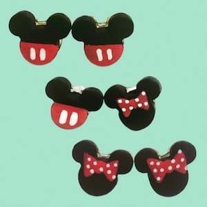 May include: Six pairs of black and red Mickey Mouse and Minnie Mouse-themed earrings. The Mickey Mouse earrings have a black head with a red face and white accents. The Minnie Mouse earrings have a red bow with white polka dots.