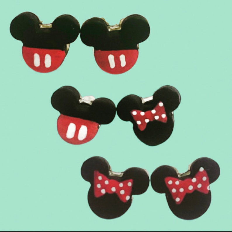 May include: Six pairs of black and red Mickey Mouse and Minnie Mouse-themed earrings. The Mickey Mouse earrings have a black head with a red face and white accents. The Minnie Mouse earrings have a red bow with white polka dots.
