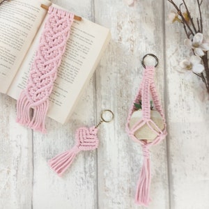 May include: Three pink macrame items: a bookmark, a keychain, and a hanging planter. The bookmark is on a book, the keychain is on a white wooden surface, and the planter is hanging from a key ring.