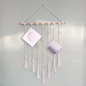 Könnte beinhalten: Ein weißer Makramee-Wandbehang mit Holzleiste und Quasten. Zwei weiße Karten werden mit Bildern von Blumen und einer Landschaft angezeigt.