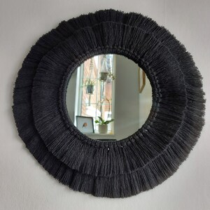 Puede incluir: Un espejo redondo con un borde de flecos negros. El espejo refleja una ventana con plantas.