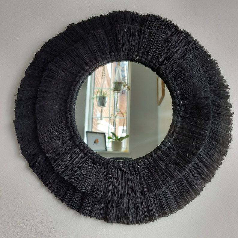 Puede incluir: Un espejo redondo con un borde de flecos negros. El espejo refleja una ventana con plantas.