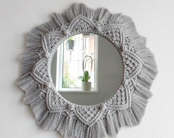 May include: A gray macrame mirror with a round mirror and a fringe border. The mirror is reflecting a window with a plant in it.