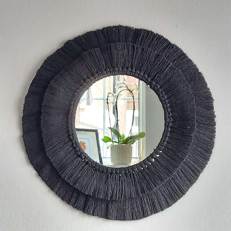Puede incluir: Un espejo redondo negro con un marco grueso y con flecos. El espejo refleja una ventana con una planta en maceta.