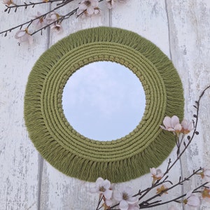 May include: A round mirror with a green macrame frame and fringe. The mirror is surrounded by a thick, textured frame with a fringe border.