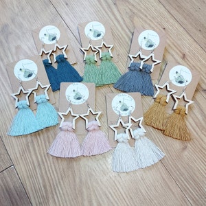Op de afbeelding: Assortiment van handgemaakte stervormige oorbellen met kleurrijke kwastjes. De oorbellen zijn gemaakt van hout en zijn verkrijgbaar in verschillende kleuren, waaronder roze, blauw, groen en bruin. De oorbellen worden gepresenteerd op een houten oppervlak.