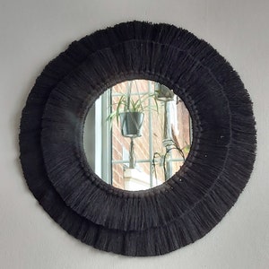 Puede incluir: Un espejo redondo con un marco de flecos negros. El espejo refleja una ventana con una planta en maceta.
