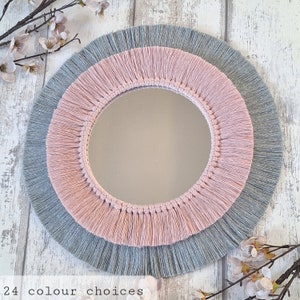 May include: A round mirror with a grey and pink fringe border. The mirror is framed with a thick, layered fringe that is tied together with a knot at the top.