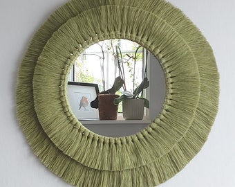 Puede incluir: Un espejo redondo con un borde de flecos verdes. El espejo refleja una ventana con plantas y un cuadro enmarcado.