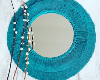 Puede incluir: Un espejo redondo de macramé turquesa con un diseño de flecos. El marco del espejo está hecho de macramé en capas, creando un borde decorativo texturizado. El espejo es circular y refleja el entorno. Ramas decorativas con capullos blancos en primer plano.