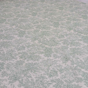 May include: A close-up of a white fabric with a green floral pattern. The fabric has a subtle texture and a delicate design.