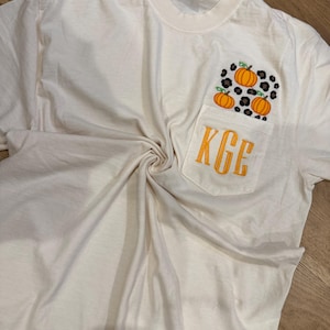 Könnte beinhalten: Ein cremefarbenes T-Shirt mit einer Brusttasche, die ein Leopardenmuster, drei orangefarbene Kürbisse mit grünen Stielen und die gelb gestickten Monogrammbuchstaben KGE auf der Tasche zeigt.