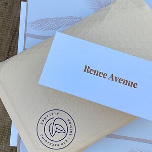 May include: A beige bubble mailer with a circular Noissue Eco Packaging Alliance logo in dark blue, topped with a white rectangular card featuring the brown text Renee Avenue, resting on a white box with a light grey palm tree pattern.