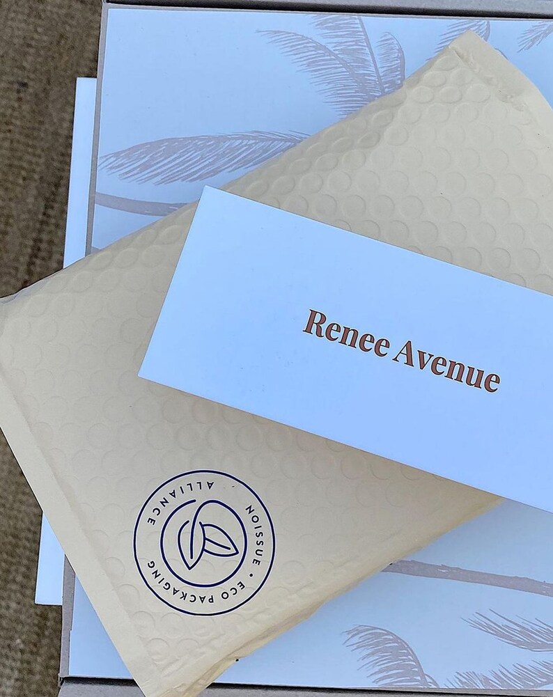 May include: A beige bubble mailer with a circular Noissue Eco Packaging Alliance logo in dark blue, topped with a white rectangular card featuring the brown text Renee Avenue, resting on a white box with a light grey palm tree pattern.