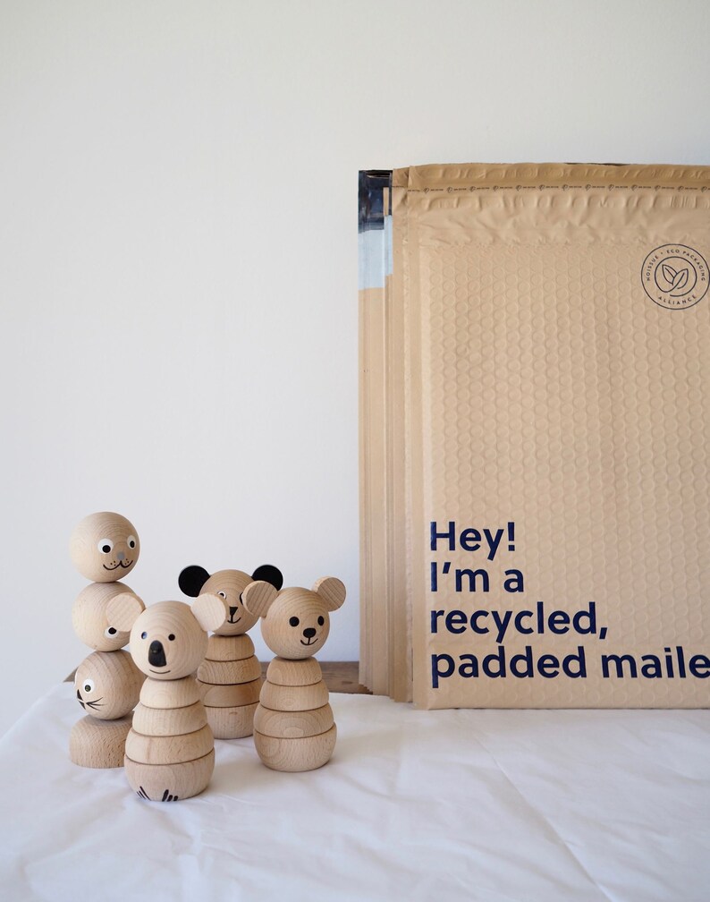 May include: Four wooden animal figurines with smooth, light-coloured surfaces and painted facial features, standing next to a stack of tan, bubble-padded mailers with the printed text: Hey! I'm a recycled, padded mailer.