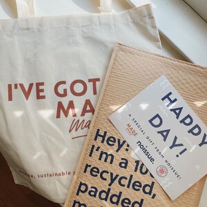 May include: A cream-coloured canvas tote bag with the text I'VE GOT MA printed in brown, alongside a tan padded mailer with the text Hey: I'm a 100% recycled, padded mailer. A white card with the text Happy Day! A SPECIAL GIFT FROM NOISSUE! is included.