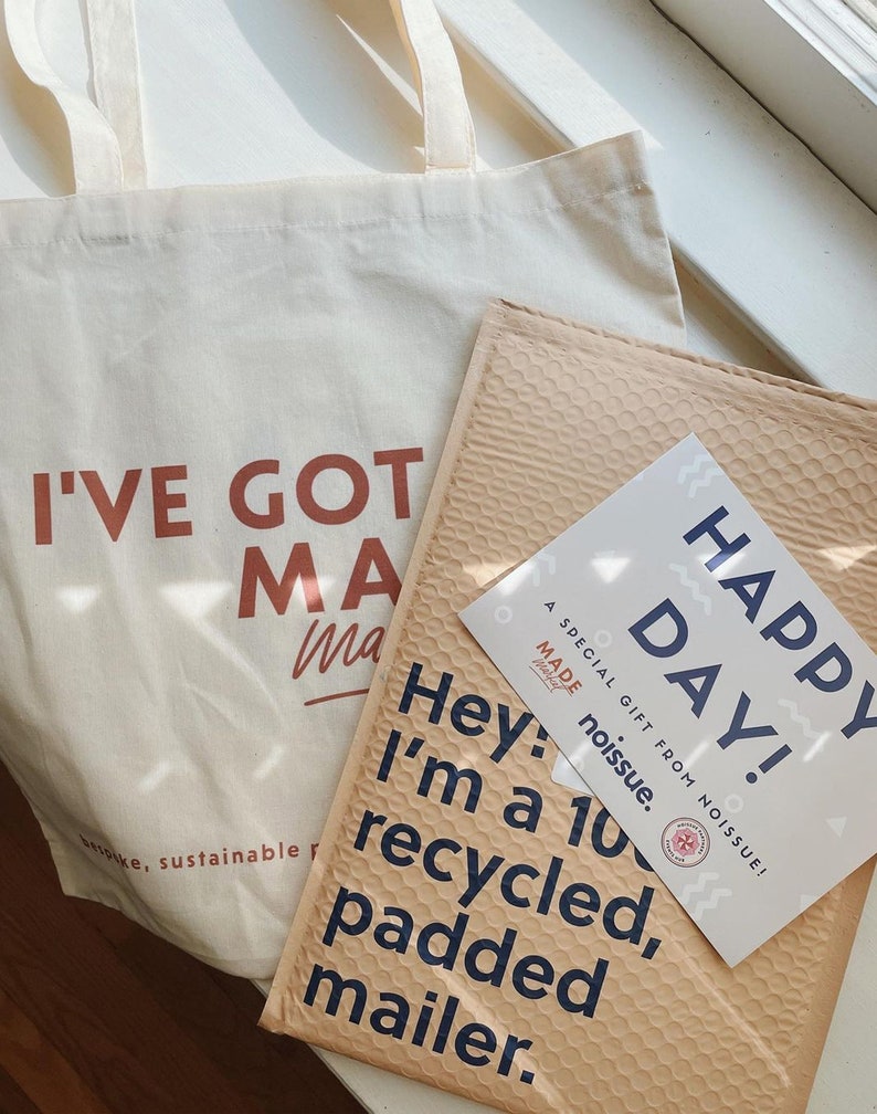 May include: A cream-coloured canvas tote bag with the text I'VE GOT MA printed in brown, alongside a tan padded mailer with the text Hey: I'm a 100% recycled, padded mailer. A white card with the text Happy Day! A SPECIAL GIFT FROM NOISSUE! is included.
