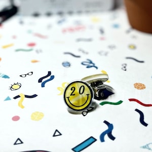 May include: A pair of cufflinks with a yellow smiley face design. The smiley face has the number 20 inside it. The cufflinks are on a white background with colorful confetti.