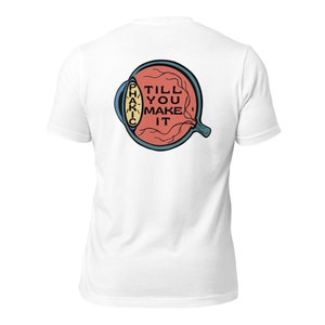 以下が含まれることがあります： 目のグラフィックが描かれた白いTシャツ。目の中に「TILL YOU MAKE IT」という文字があります。