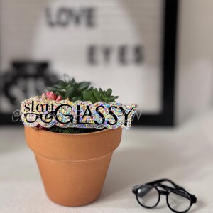 Puede incluir: Una pequeña maceta de terracota con una planta suculenta y una cubierta brillante "slay glassy". Un par de gafas con montura negra descansa cerca. El fondo presenta un letrero que dice "Eye Love Eyes".