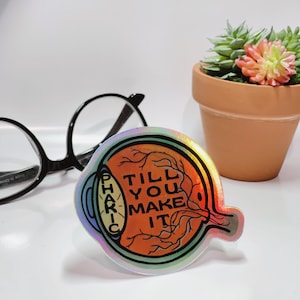 May include: A holographic sticker with an eye illustration and the text "TILL YOU MAKE IT" inside the eye. The eye is surrounded by a black outline and has a red and orange color scheme.