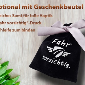 May include: A black velvet gift bag with a white ribbon and the text "Fahr vorsichtig." printed on the front. The bag is on a wooden surface.