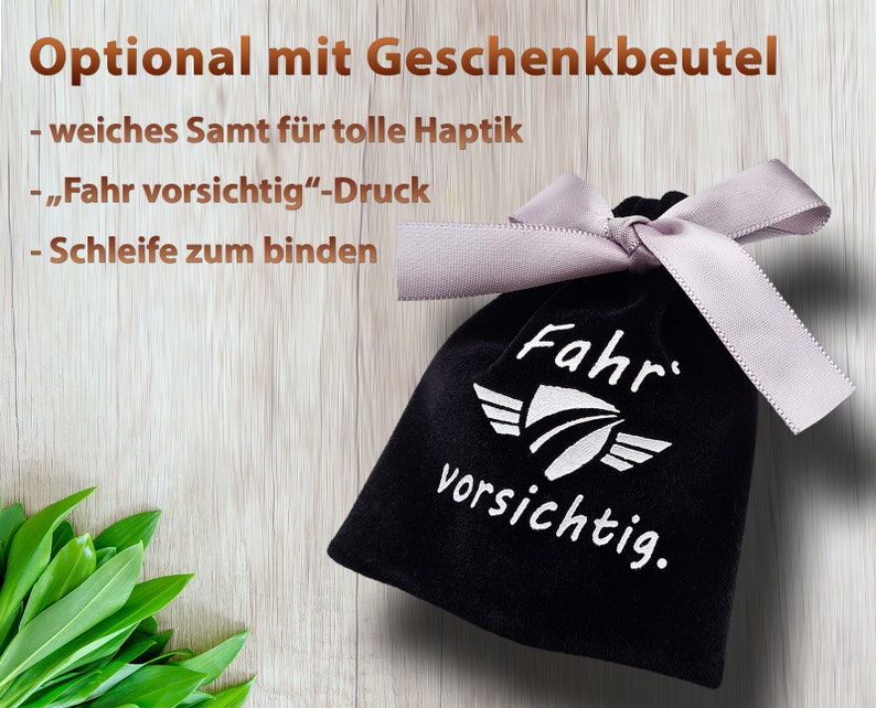 May include: A black velvet gift bag with a white ribbon and the text "Fahr vorsichtig." printed on the front. The bag is on a wooden surface.