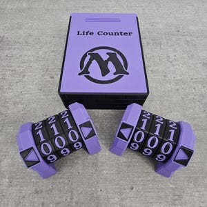 May include: A purple rectangular box labeled Life Counter with a black logo, accompanied by two purple mechanical life counter devices. Each device features three rotating number wheels displaying digits from zero to two and black arrow indicators.