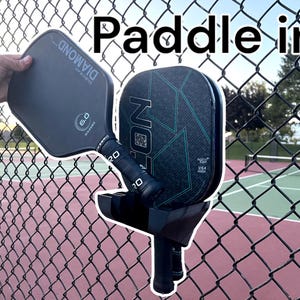 May include: Two black and green pickleball paddles are mounted on a black metal rack. The rack is attached to a chain link fence. The paddles are both new and have the brand name "Onix" printed on them. The text "Paddle in!" is written in white letters on a black background above the paddles.