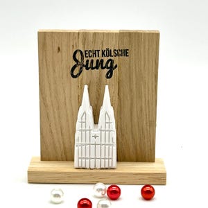 Könnte beinhalten: Ein Holzschild mit dem Text "Echt Kölsche Jung" und einem weißen Modell des Kölner Doms. Das Schild steht auf einem Holzsockel und es liegen rote und weiße Perlen herum.