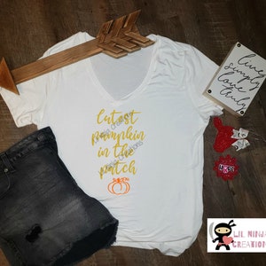 Puede incluir: Camiseta blanca con cuello en V con el texto dorado "Cutest Pumpkin in the Patch" y un gráfico de calabaza naranja. La camiseta se combina con pantalones cortos de mezclilla negros y una decoración de flecha de madera.