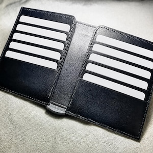Peut inclure: Un portefeuille en cuir noir ouvert à plat, présentant plusieurs fentes horizontales pour cartes sur les côtés gauche et droit. Le portefeuille contient plusieurs cartes rectangulaires blanches vierges insérées dans les fentes, avec une charnière métallique visible au centre.