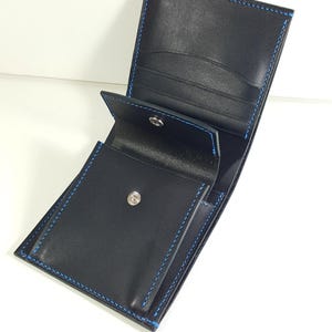May include: A black leather bifold wallet featuring vibrant blue stitching along the edges. The interior includes multiple card slots and a central coin pocket secured with a silver-toned metal snap button closure.