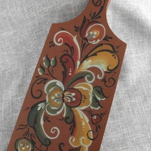 May include: A wooden cutting board with a handle, painted with a floral design in shades of brown, yellow, red, and green. The decorative board features a detailed, hand-painted floral pattern, ideal for kitchen decor.