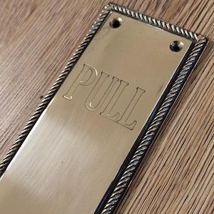 Könnte beinhalten: Rechteckige Messing-Türzugplatte mit dem Wort „PULL“ in der Mitte eingraviert. Die Platte hat einen seilartigen Rand und zwei Schraubenlöcher oben. Die Oberfläche ist goldfarben glänzend.