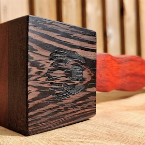 May include: A wooden mallet with a dark brown and red wood handle and a square head made of dark wood with a black and white design. The head of the mallet has a carved design of a stylized bird.
