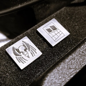 Puede incluir: Dos gemelos cuadrados de plata con diseños grabados. Uno presenta una calavera alada, el otro un patrón geométrico. Se encuentran sobre una superficie oscura y texturizada, mostrando los detalles.