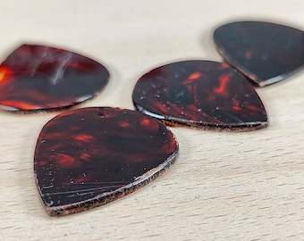 May include: Four tortoise shell guitar picks on a wooden surface. The picks are all different shapes and sizes, but they are all made of the same material.