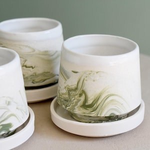 May include: A collection of white ceramic planters with a marbled green design. Each planter sits on a matching white saucer. The planters have a cylindrical shape with a slightly flared top, and the green marbling adds a unique touch.