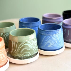 May include: A collection of ceramic planters in various colors, including blue, green, purple, and black. Each planter features a unique marbled design and sits on a white saucer. The planters are suitable for small plants.