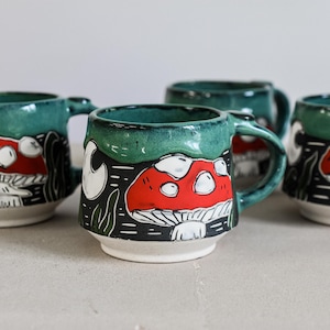 May include: A set of four ceramic mugs featuring a hand-painted design of red mushrooms with white spots on a black background. The upper portion of each mug is glazed in a deep teal green, while the base is a contrasting off-white ceramic.