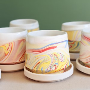 May include: A collection of ceramic planters with a white base and a colorful, marbled design in shades of yellow, red, and blue. Each planter sits on a matching saucer. The background is a solid green.
