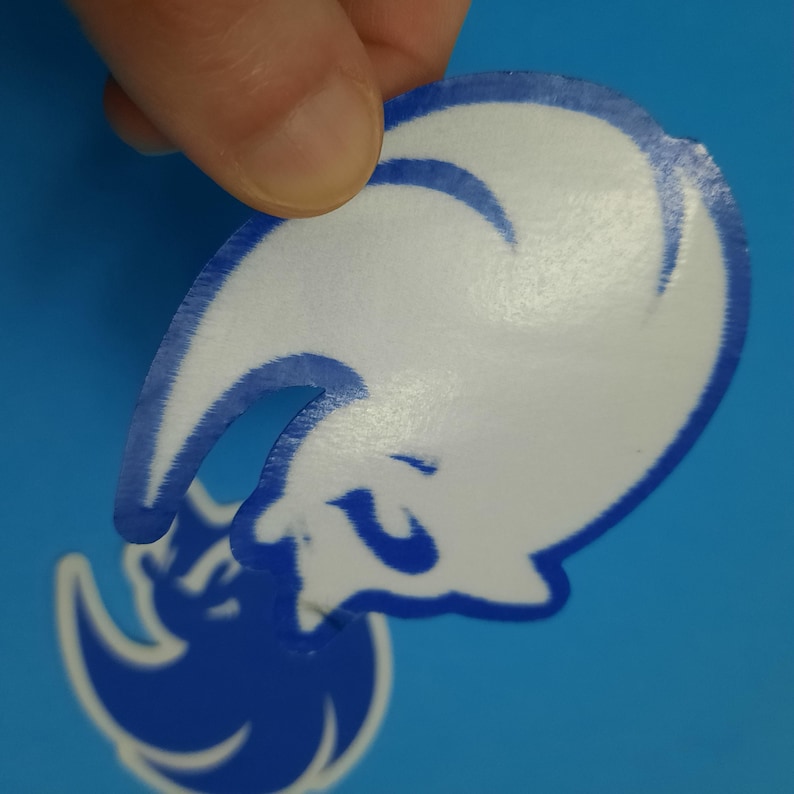 May include: A white sticker with a blue outline and a stylized design, held by a person's fingers. The sticker is on a blue background. The design appears to be a stylized animal.