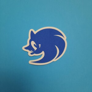 May include: A blue and white patch featuring the head of Sonic the Hedgehog. The character's face is in profile, with a smiling expression. The patch has a white border and is set against a light blue background.